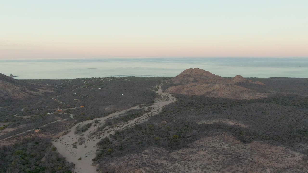 Aerial view in Cabo Pulmo with the dirt road running diagonally through arid vegetation, a rounded rocky ridge in the midground, the ocean in back, and no visible people.