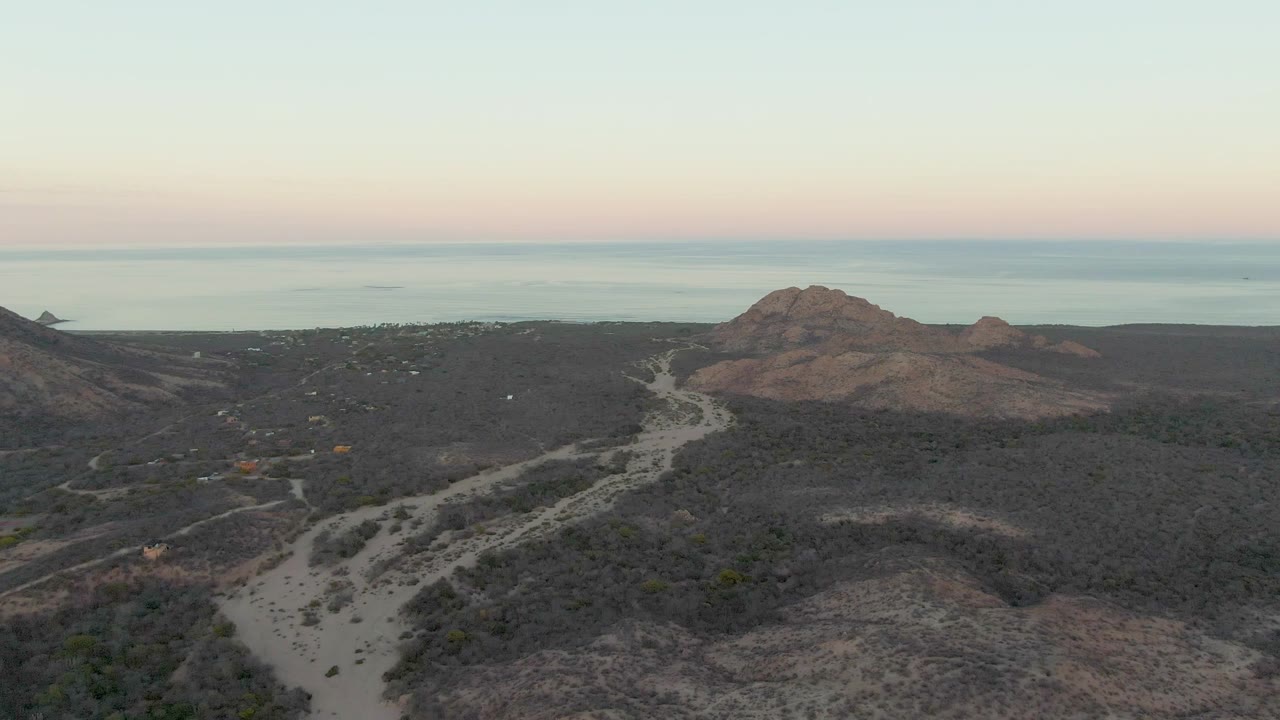 Wide drone frame over Cabo Pulmo terrain, showing a winding pale road, dark scrub-covered hills, rocky outcrops, and a smooth ocean horizon beneath pink and blue dawn color, with no visible people.