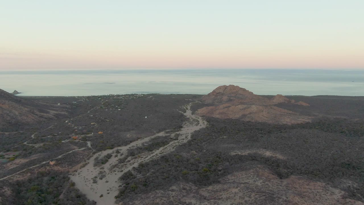 Cabo Pulmo aerial frame with the dirt road bending through dry hilly terrain, a rounded hill on the right, calm sea beyond, and no visible people.