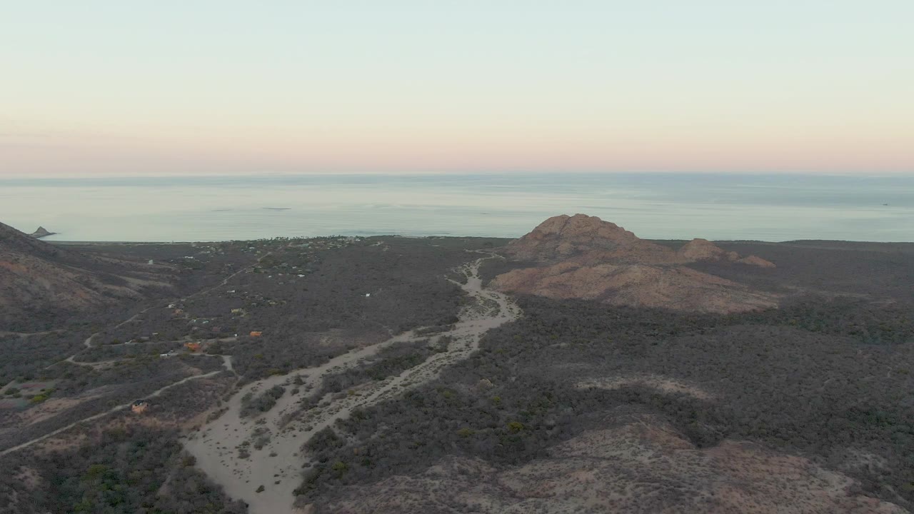 Aerial still from Cabo Pulmo showing a winding dirt track in the foreground, scrub-covered slopes, a rocky hill near the right side, the ocean horizon, and no visible people.