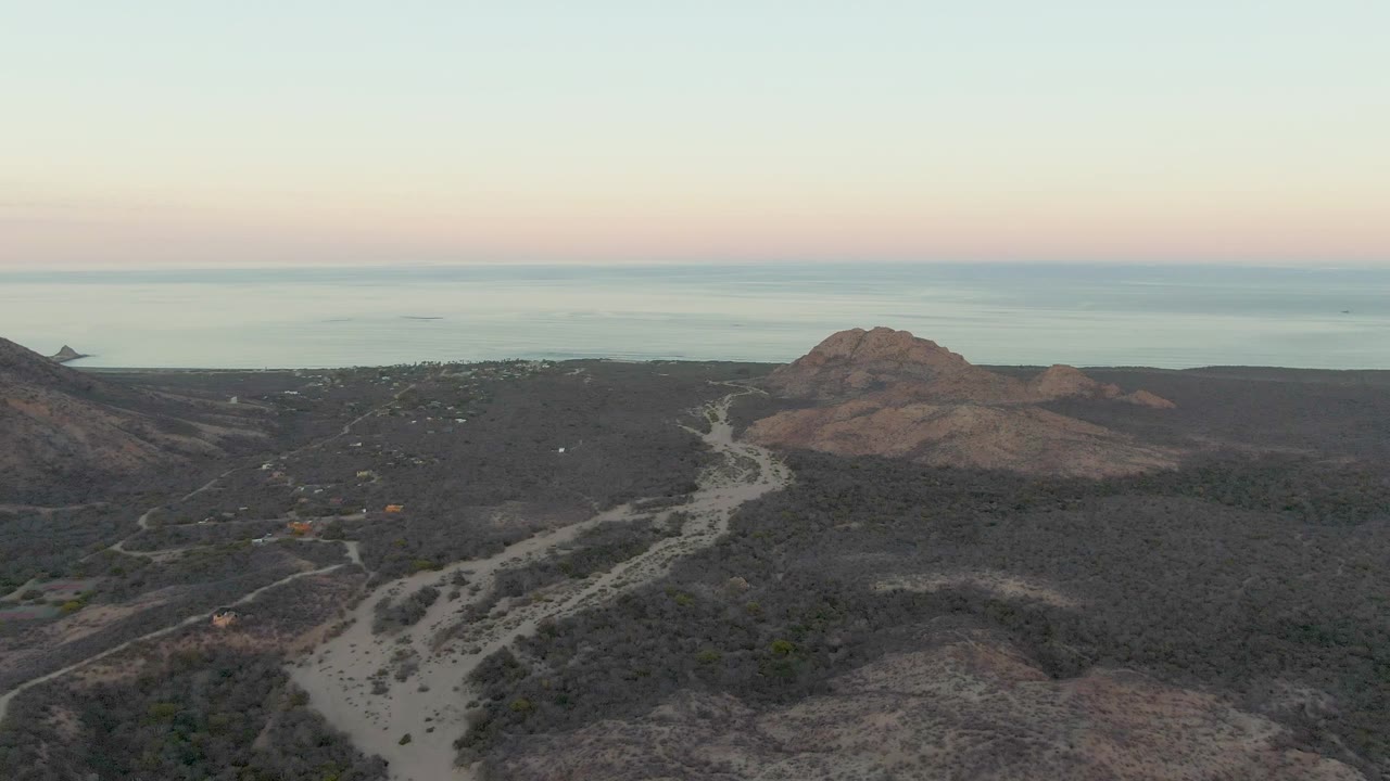 Wide drone-style view of Cabo Pulmo coastal hills, with a pale road threading through darker brush, a rounded hill to the right, calm ocean in the distance, and no visible people.