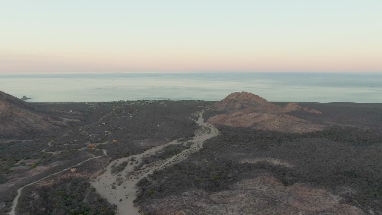 Opening aerial frame over Cabo Pulmo dry hills, with a pale winding dirt road below, the ocean beyond the ridgeline, a pink-blue dusk sky, and no visible people.