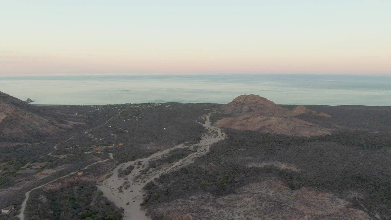 Wide drone view of a dirt road crossing arid hills with ocean in the background.