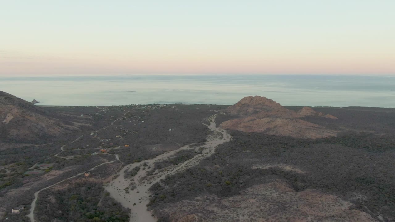 Mid-clip aerial frame over Cabo Pulmo with a winding pale road, dark hillside ridges, scattered brush, distant ocean water, and no visible people.