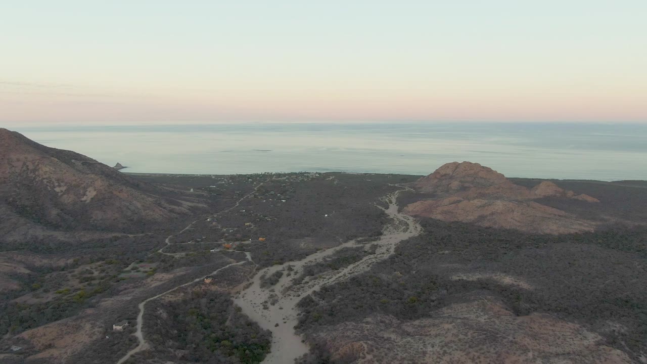 Drone perspective above Cabo Pulmo arid terrain, with the road curving through dark scrubland between a large rocky slope and a rounded hill, no visible people.