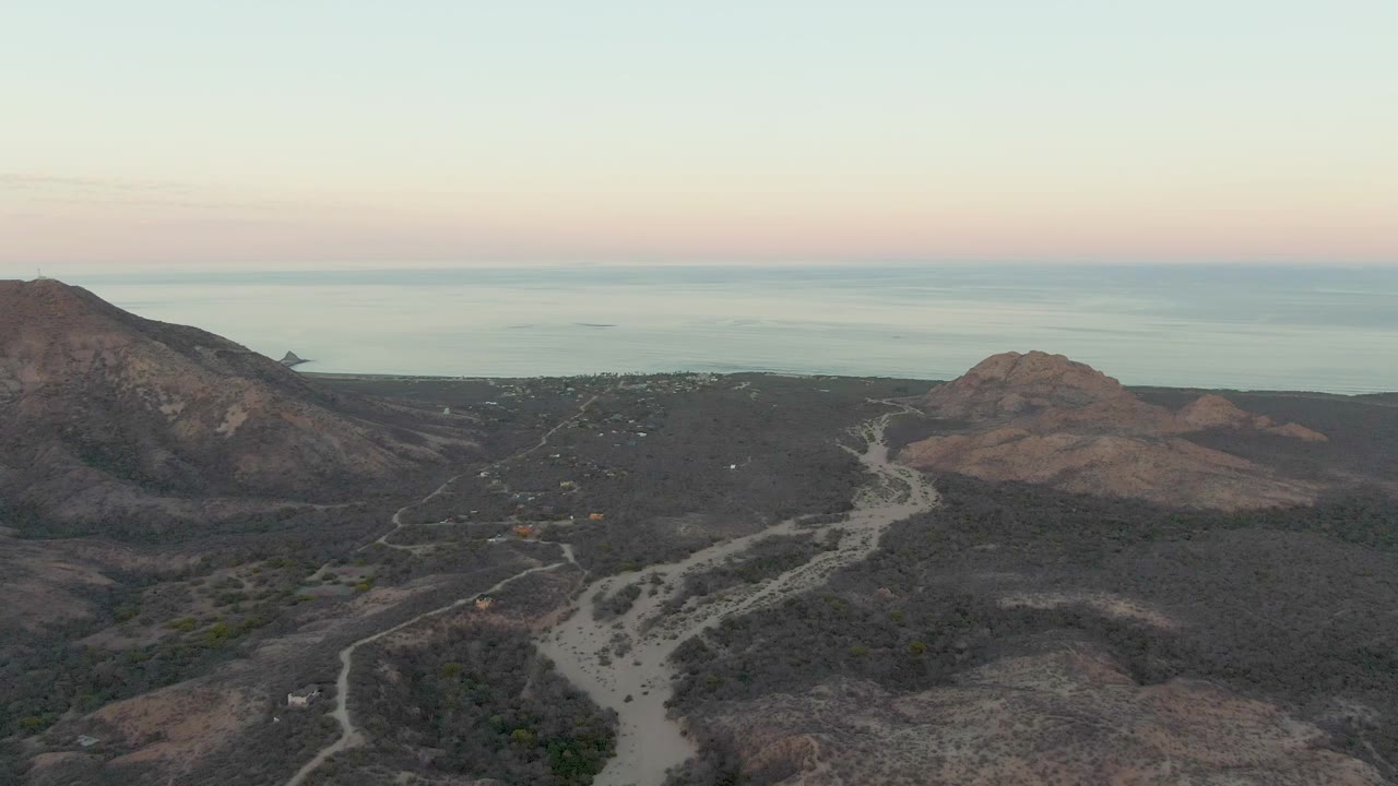 Wide aerial frame from Cabo Pulmo showing a light dirt track across brown hills, the ocean beyond the land, and a soft pink band in the sky with no visible people.