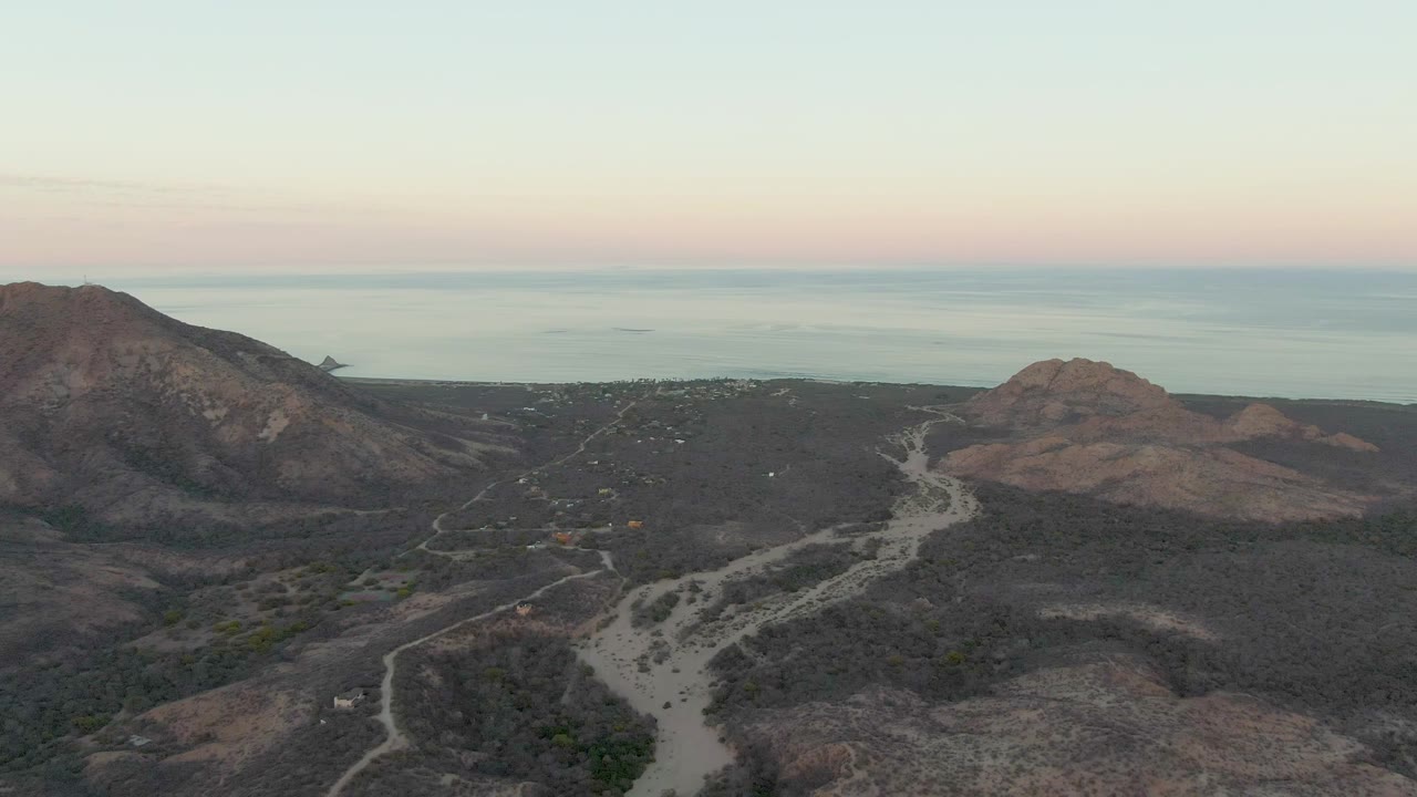 Aerial view in Cabo Pulmo with the winding road crossing the lower frame, a rocky mountain mass on the left, a rounded hill on the right, and no visible people.