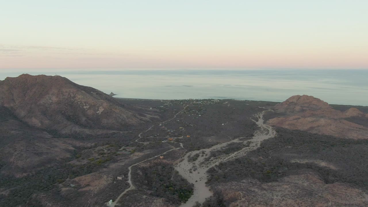 Early frame from Cabo Pulmo, Baja California Sur showing a broad aerial view of rocky hills, pale winding roads, sparse vegetation, and distant ocean with no visible people.