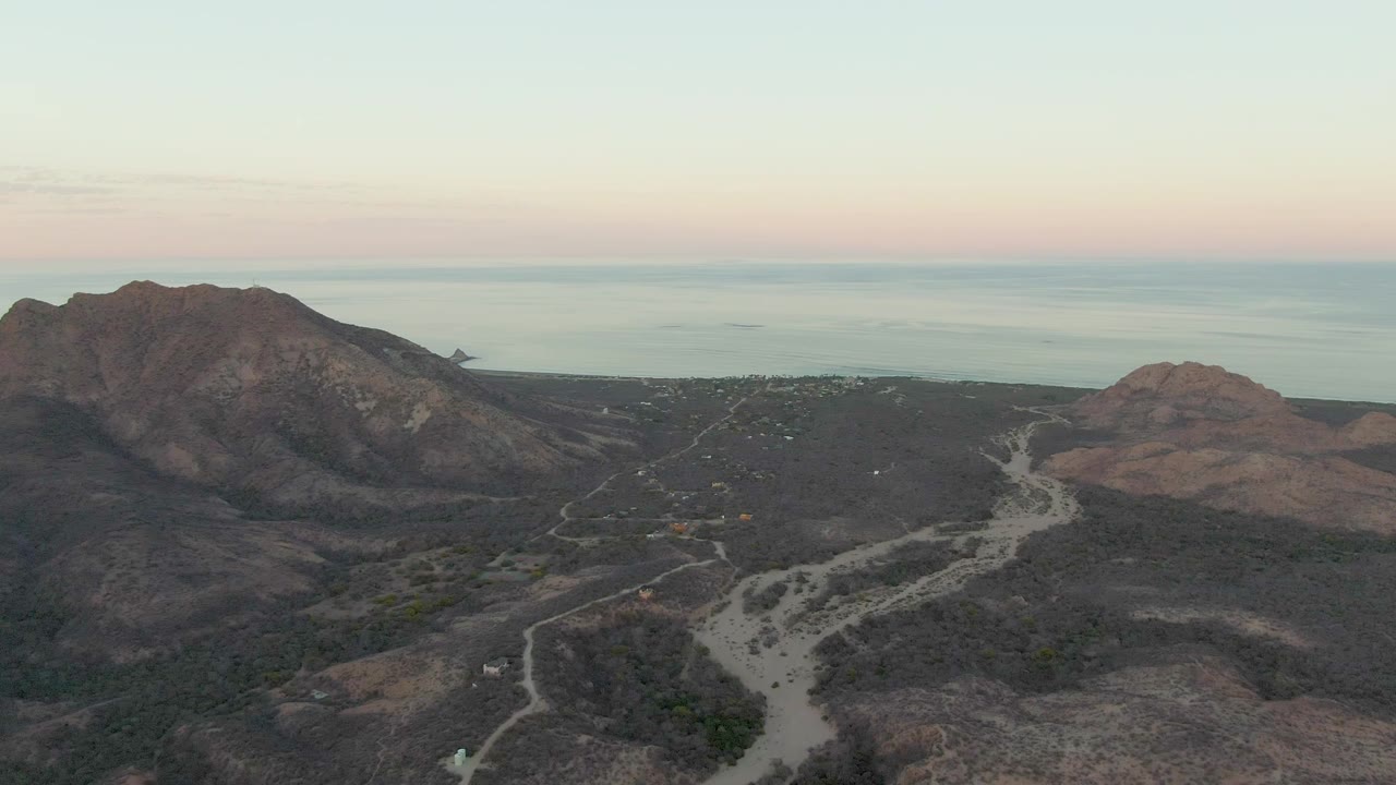 Cabo Pulmo aerial frame with the main mountain ridge shifted toward the center, pale dirt roads below, low dry vegetation, and the ocean horizon beyond with no visible people.