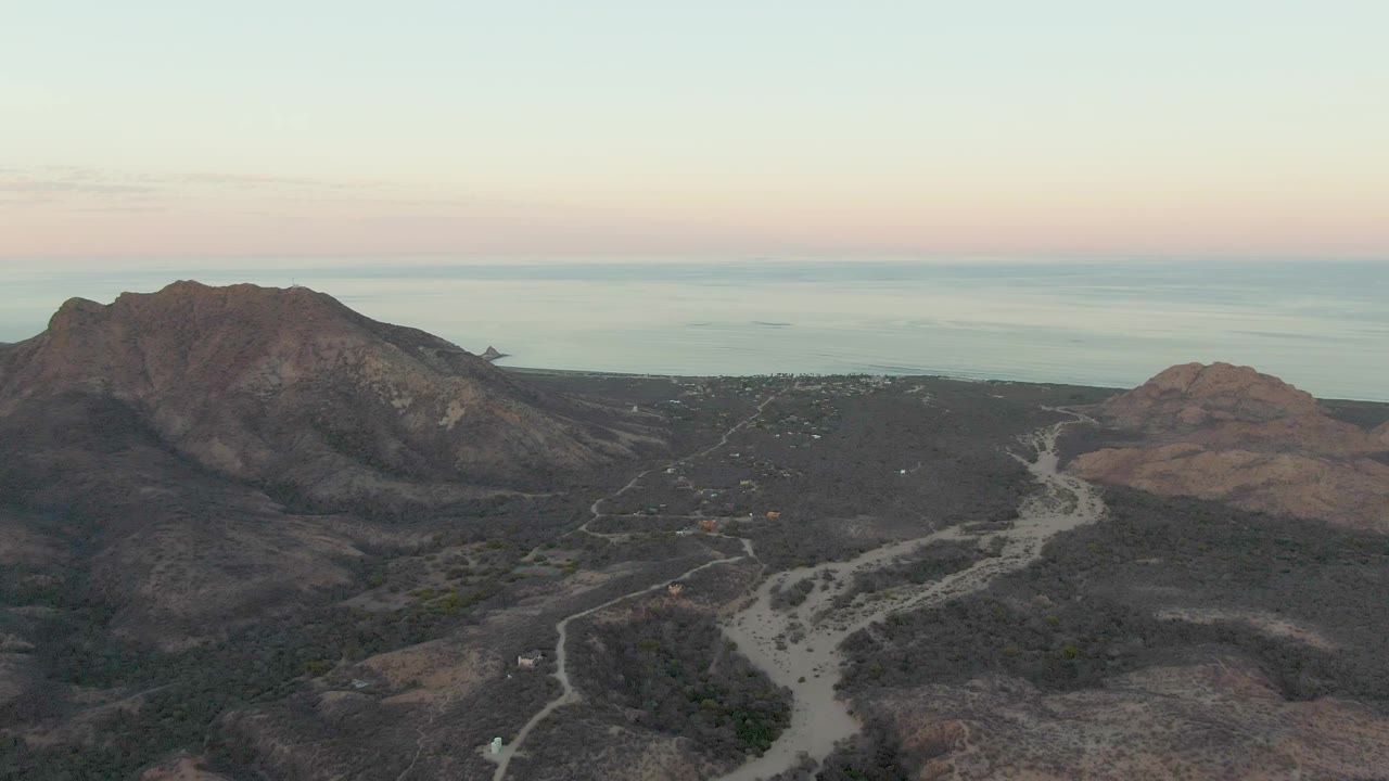 Cabo Pulmo, Baja California Sur frame showing road lines crossing dry brown terrain toward rounded hills, with a darker mountain mass and calm ocean in the distance and no visible people.