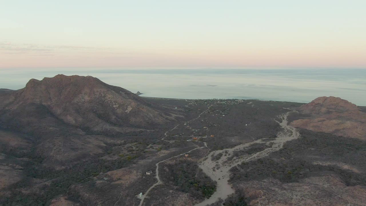 Aerial landscape with rocky hills, a winding dirt road, and calm ocean beyond.