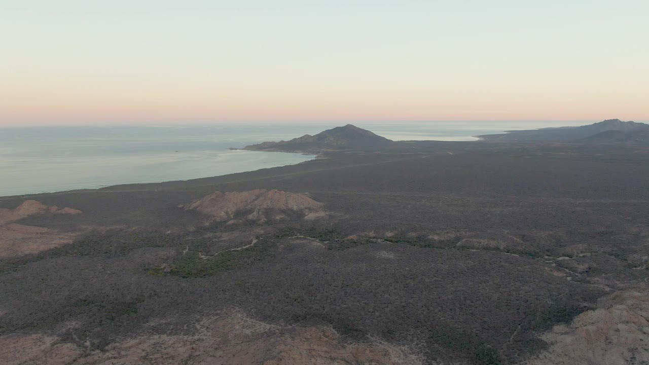 Aerial view in Cabo Pulmo keeps the rocky foreground low in frame while the headland, low hills, calm ocean, and peach-blue dusk sky remain visible, with no visible people.