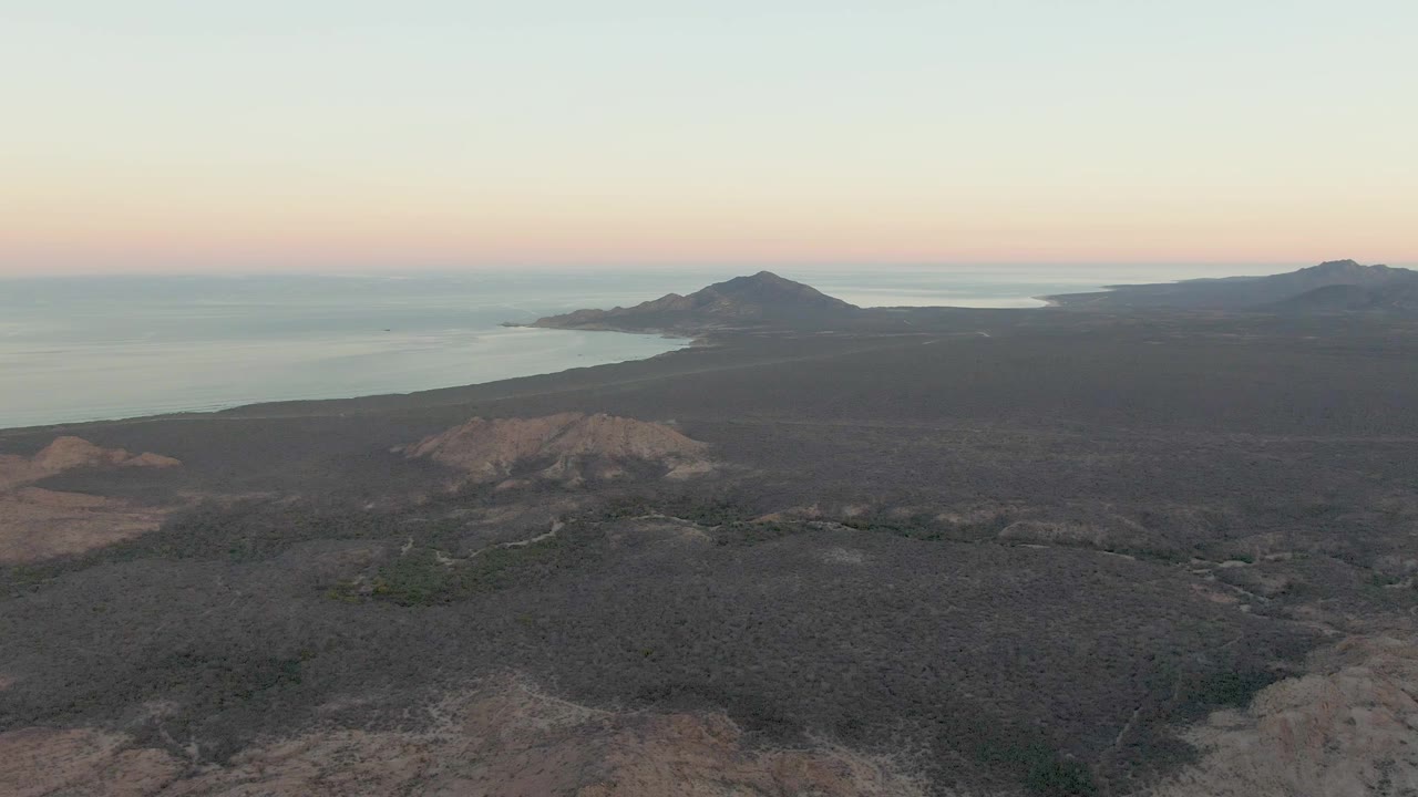 The Cabo Pulmo aerial view shows rough dark ground across the lower frame, a sloped headland near the water, distant hills, calm ocean, and a soft dusk sky, with no visible people.