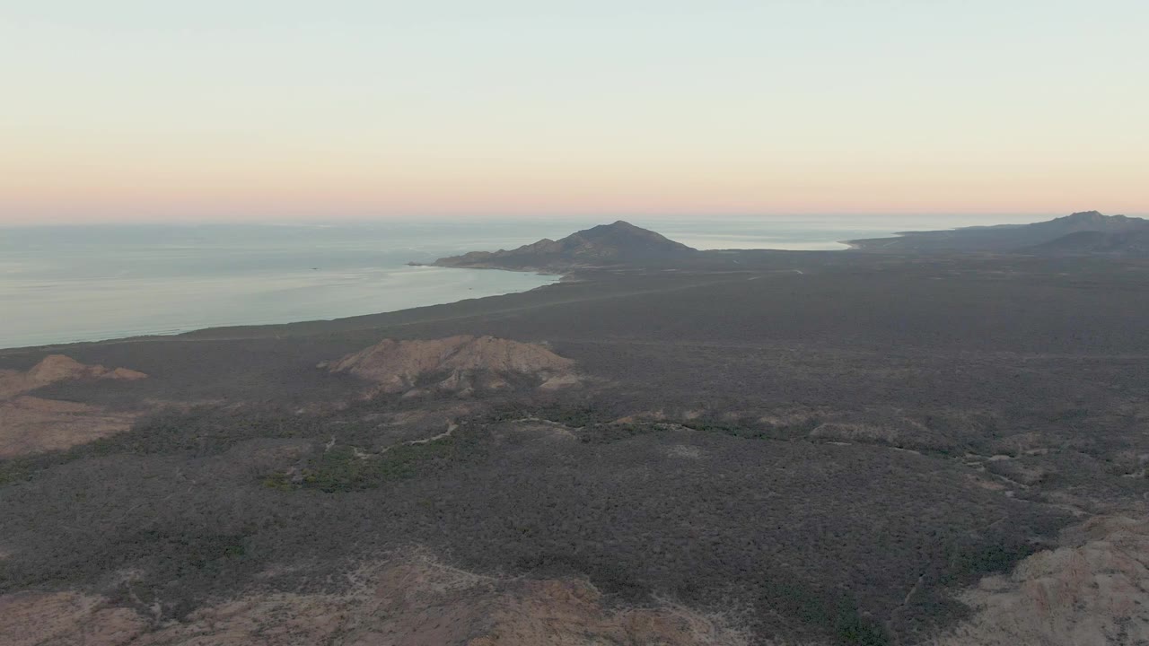 Mid-clip aerial frame over Cabo Pulmo shows arid rocky foreground, dark volcanic midground, a headland beside calm water, and a fading dusk sky with no visible people.