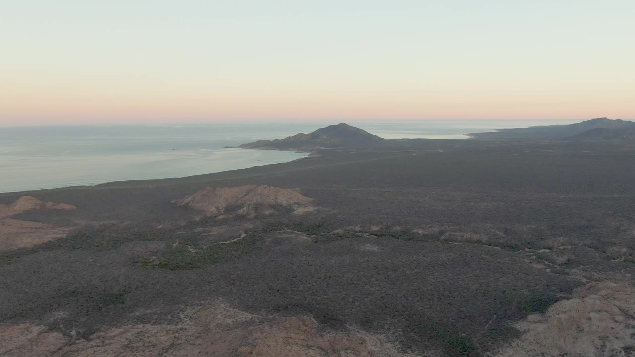 Opening frame over Cabo Pulmo volcanic coastline, with dark rocky terrain below, calm ocean to the left, distant hills on the horizon, and no visible people.