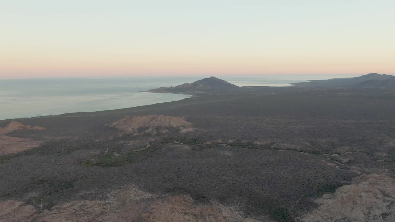 Wide aerial view of rocky coastal terrain, ocean, and a low hill under soft dusk light.