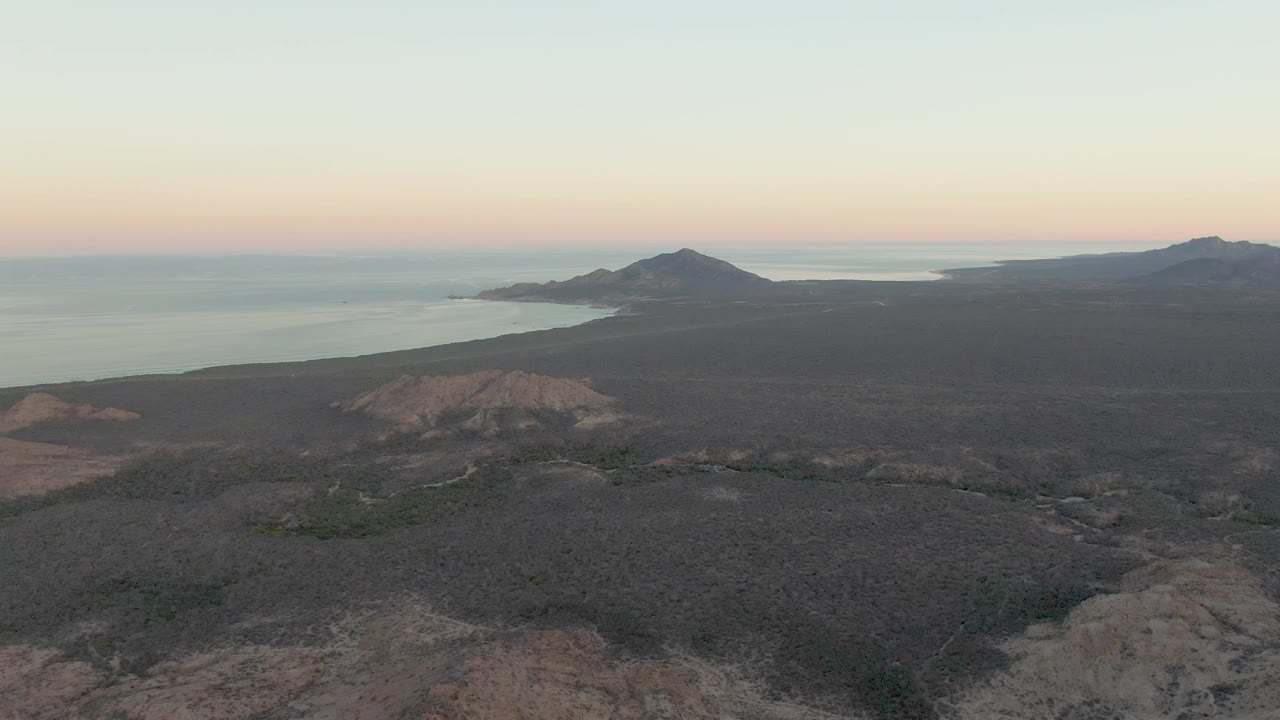 Mid-clip frame over Cabo Pulmo showing dark volcanic terrain across the foreground, calm water beyond the coast, distant hills, a pale pink-blue dusk sky, and no visible people.