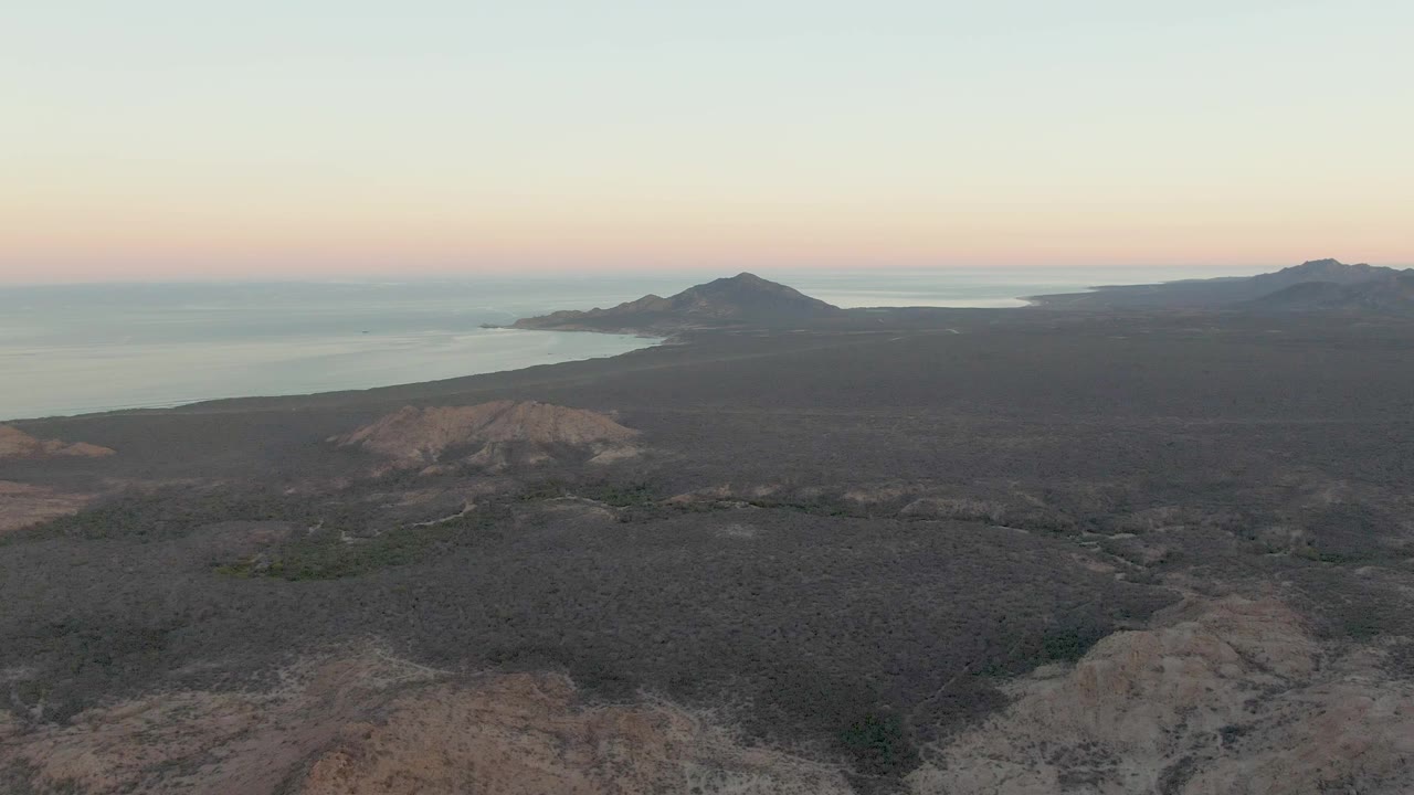 Wide aerial frame over Cabo Pulmo coastal desert with rocky foreground ridges, distant hills, calm ocean, pale dusk color, and no visible people.