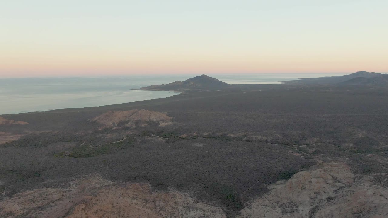 Cabo Pulmo aerial view showing a dark desert plain, rough foreground terrain, a low coastal hill, calm ocean water, and no visible people.