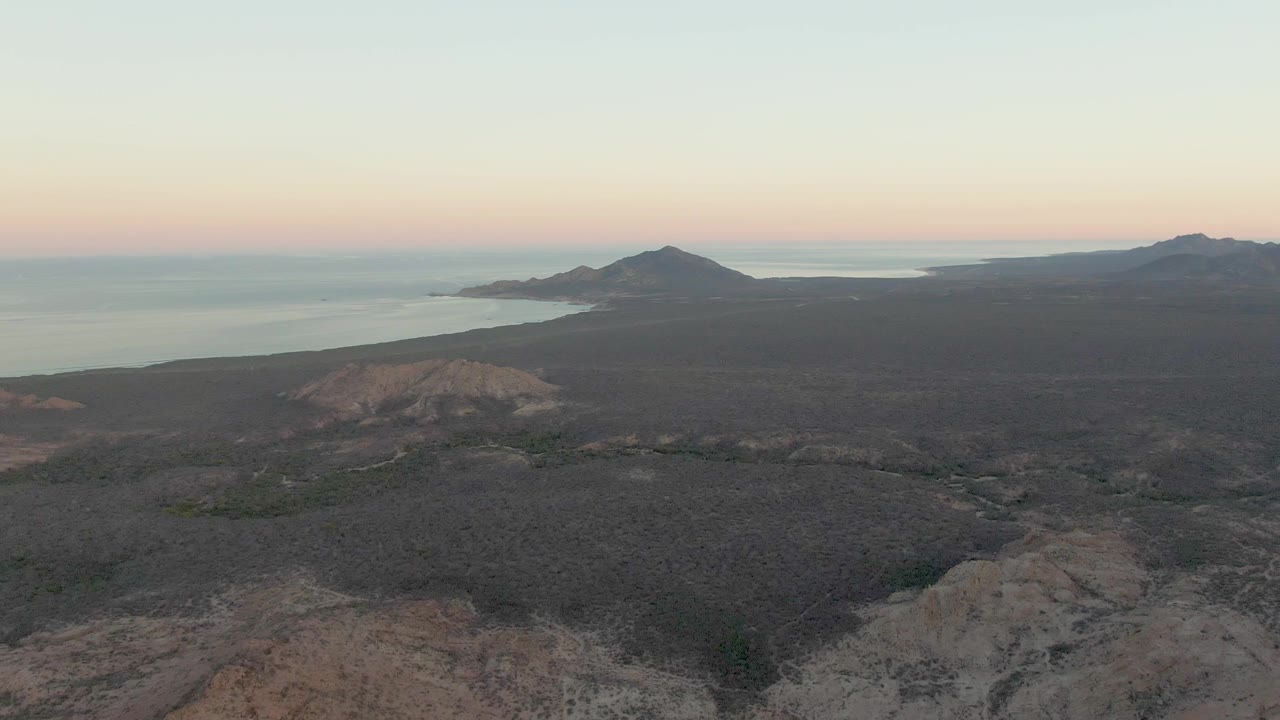 Aerial dusk frame in Cabo Pulmo with ridged dry ground in the foreground, darker scrubland across the middle, ocean behind the hills, and no visible people.