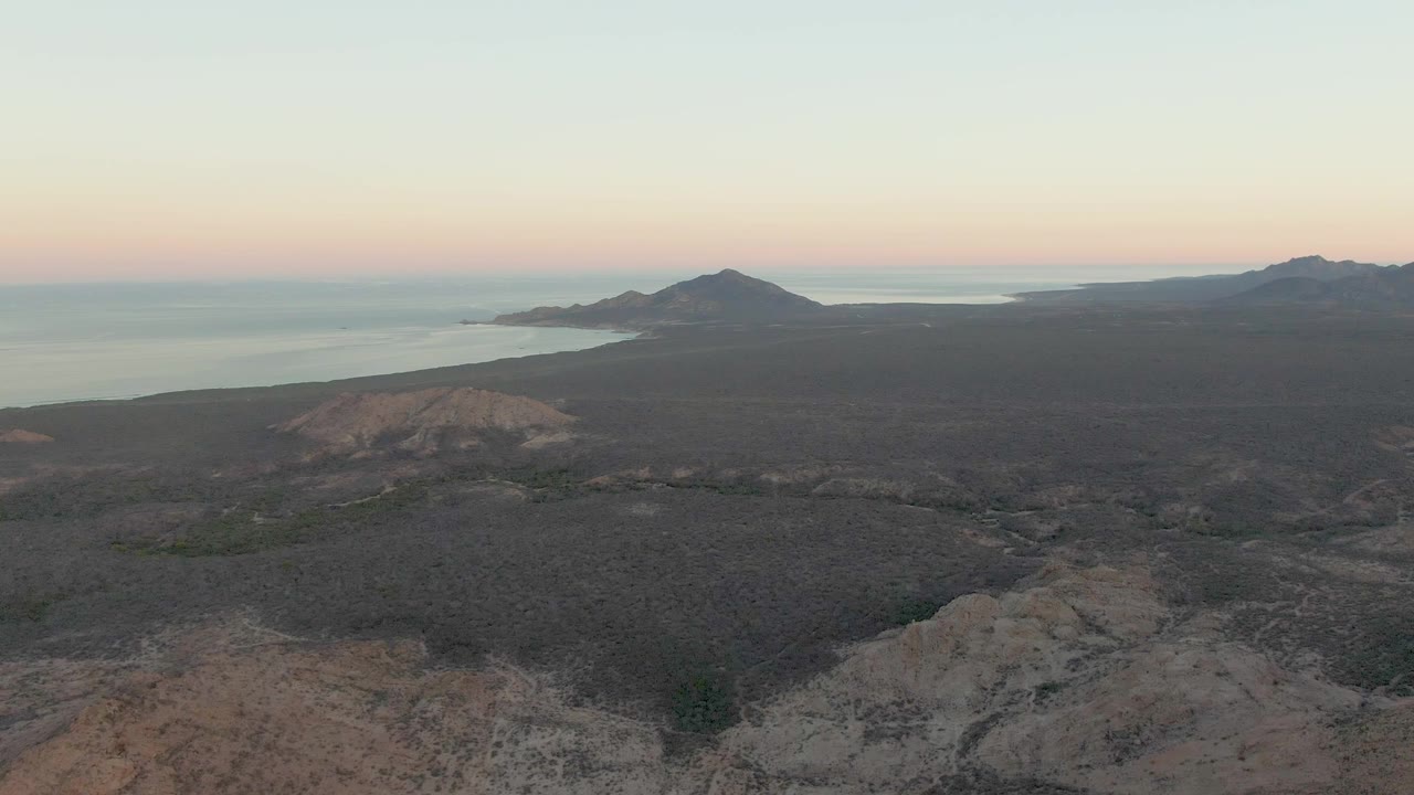 Aerial view over Cabo Pulmo volcanic terrain with pale foreground rock, dark scrubland, distant ocean, mountains on the horizon, and no visible people.