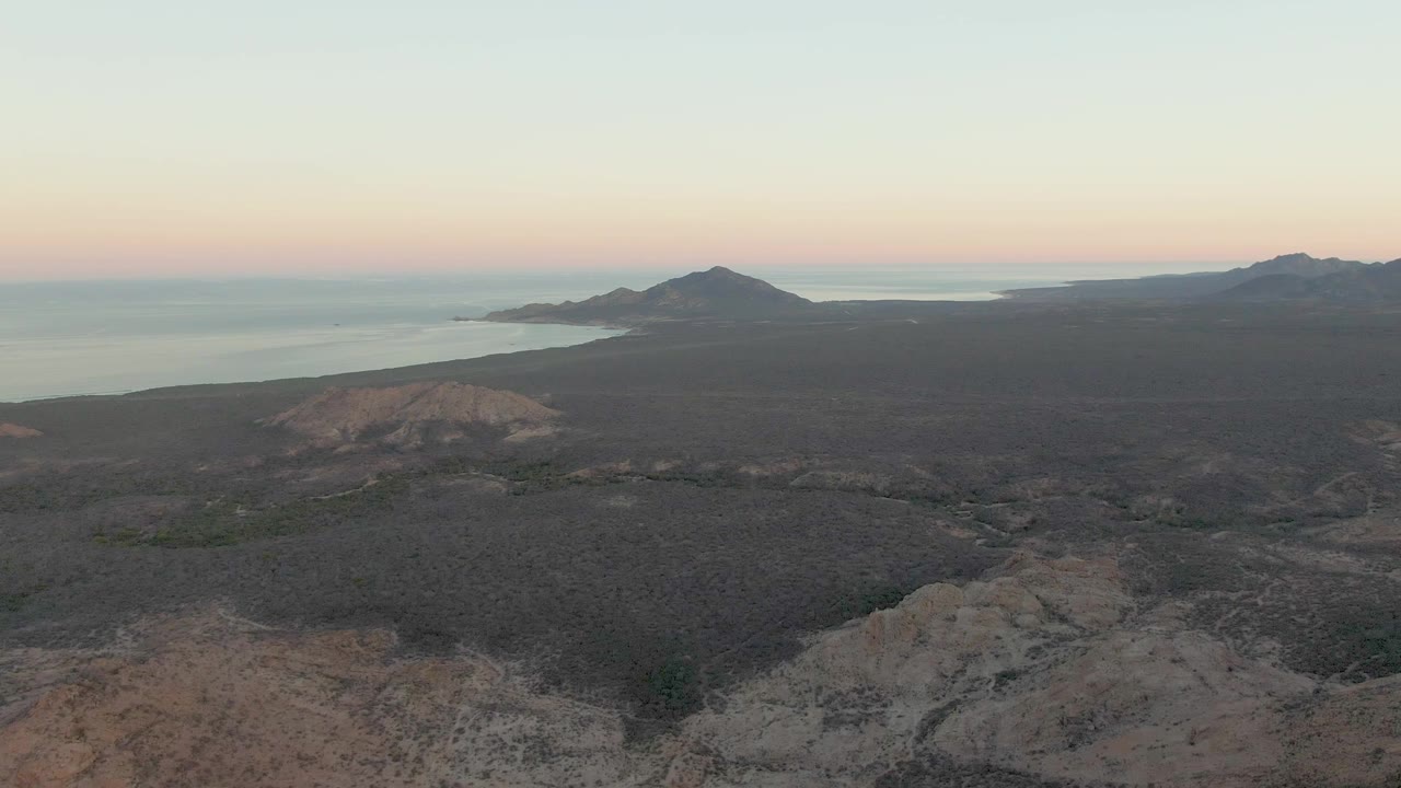 Cabo Pulmo aerial frame with foreground ridges lower in view, broad dark volcanic land, ocean beyond the coast, distant hills, and no visible people.