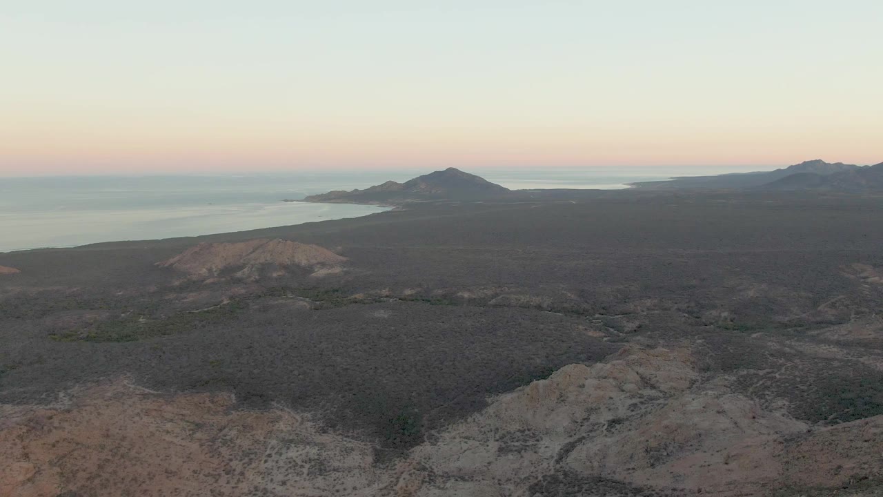 High aerial view in Cabo Pulmo showing rough volcanic foreground, a dark coastal plain, water behind the hills, a soft pink sky band, and no visible people.