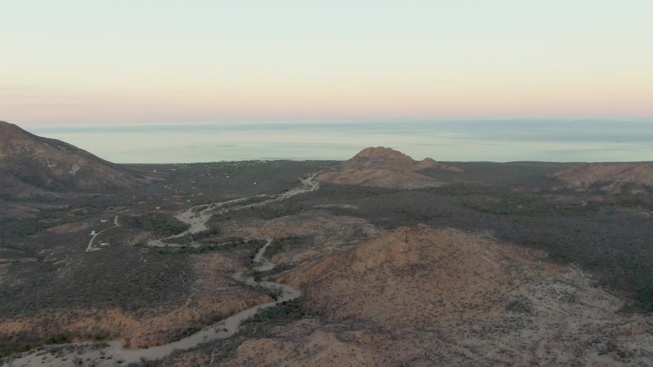 Aerial view in Cabo Pulmo, Baja California Sur with the road curving from the lower frame into dark scrub hills, a mountain slope at left, distant water, and no visible people.