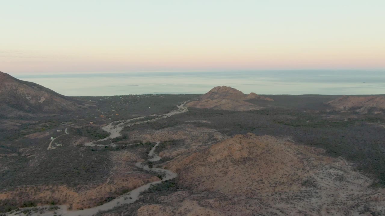 Later aerial frame from Cabo Pulmo, Baja California Sur with a broad dry foreground hill, winding road in the midground, mountains at both sides, distant water, and no visible people.