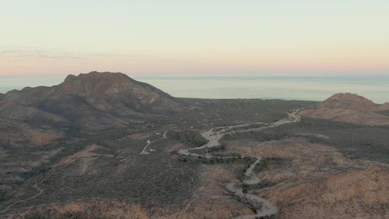 Cabo Pulmo aerial view with the pale road curving through scrub-covered dry hills, the vehicle slightly farther along the route, mountains behind it, and no visible people.