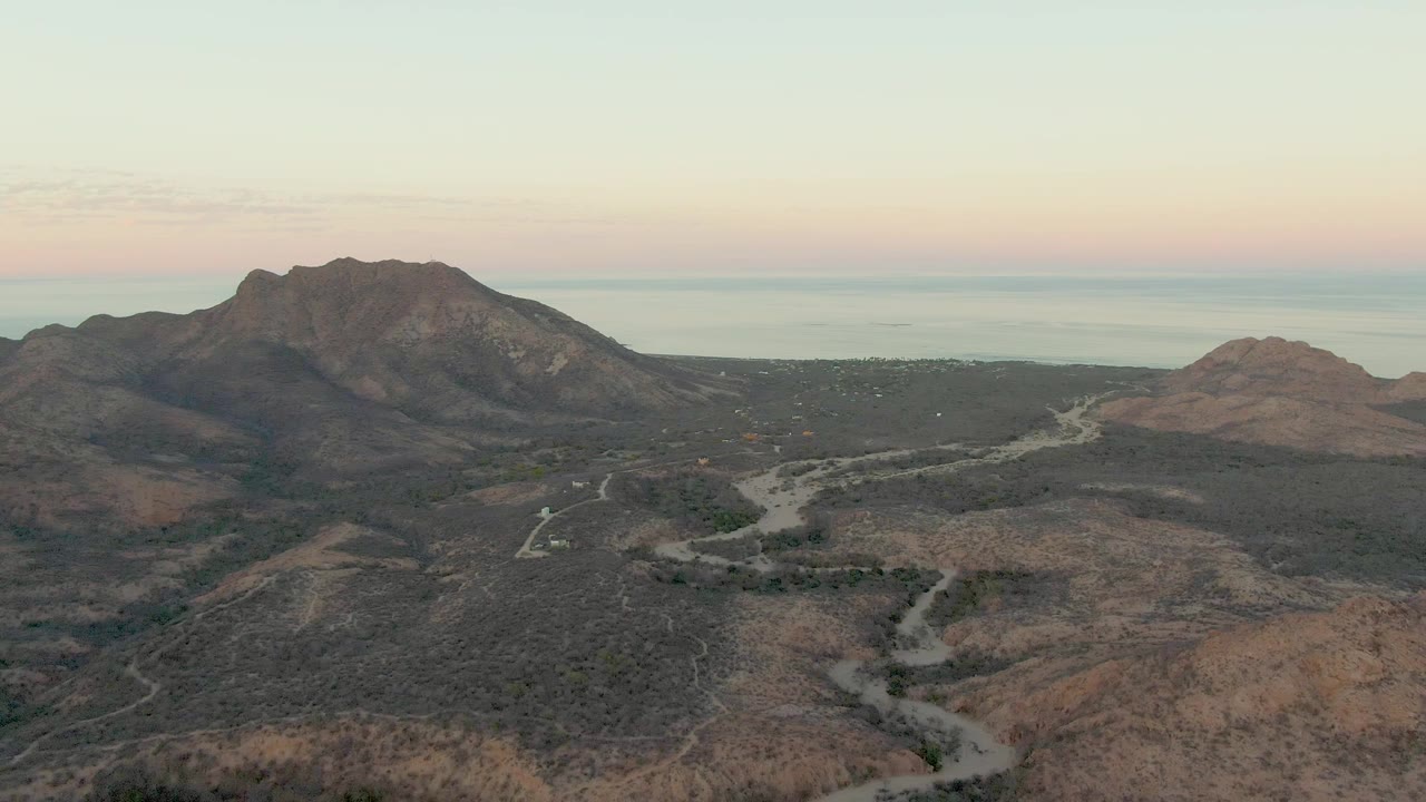 Aerial still from Cabo Pulmo with the winding dirt road running from the lower hills toward darker mountains, a small vehicle on the route, faint coastal water beyond, and no visible people.