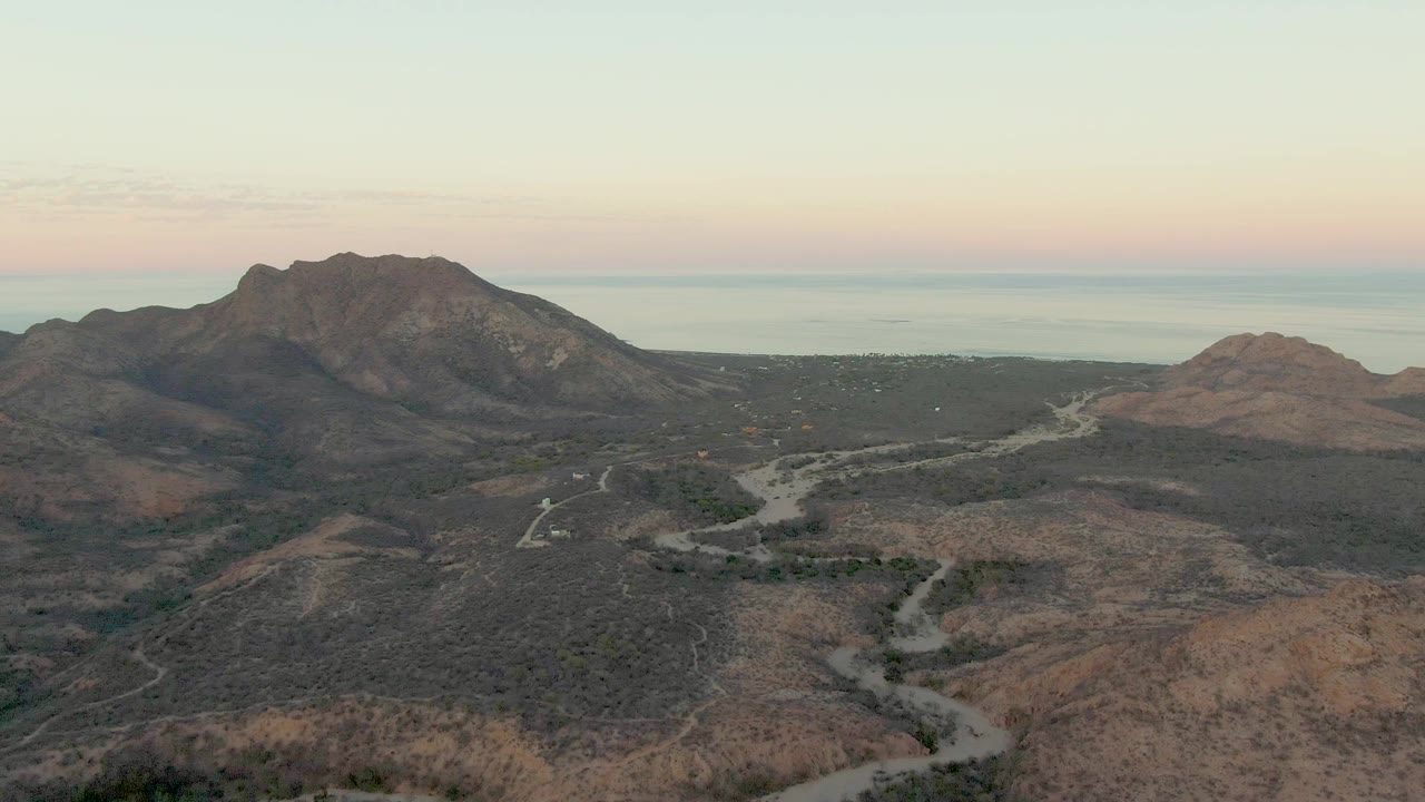 Cabo Pulmo drone frame showing the dirt road snaking through brown and gray arid hills, with the vehicle small in the scene, mountain ridges in back, water at the horizon, and no visible people.