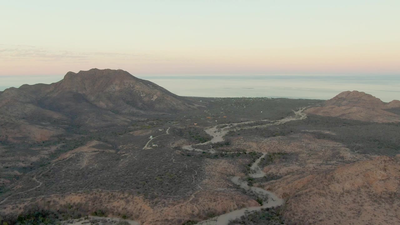 Wide aerial frame over Cabo Pulmo desert hills with winding dirt roads leading toward rocky mountains, a faint coastal horizon, warm sky color, and no visible people.