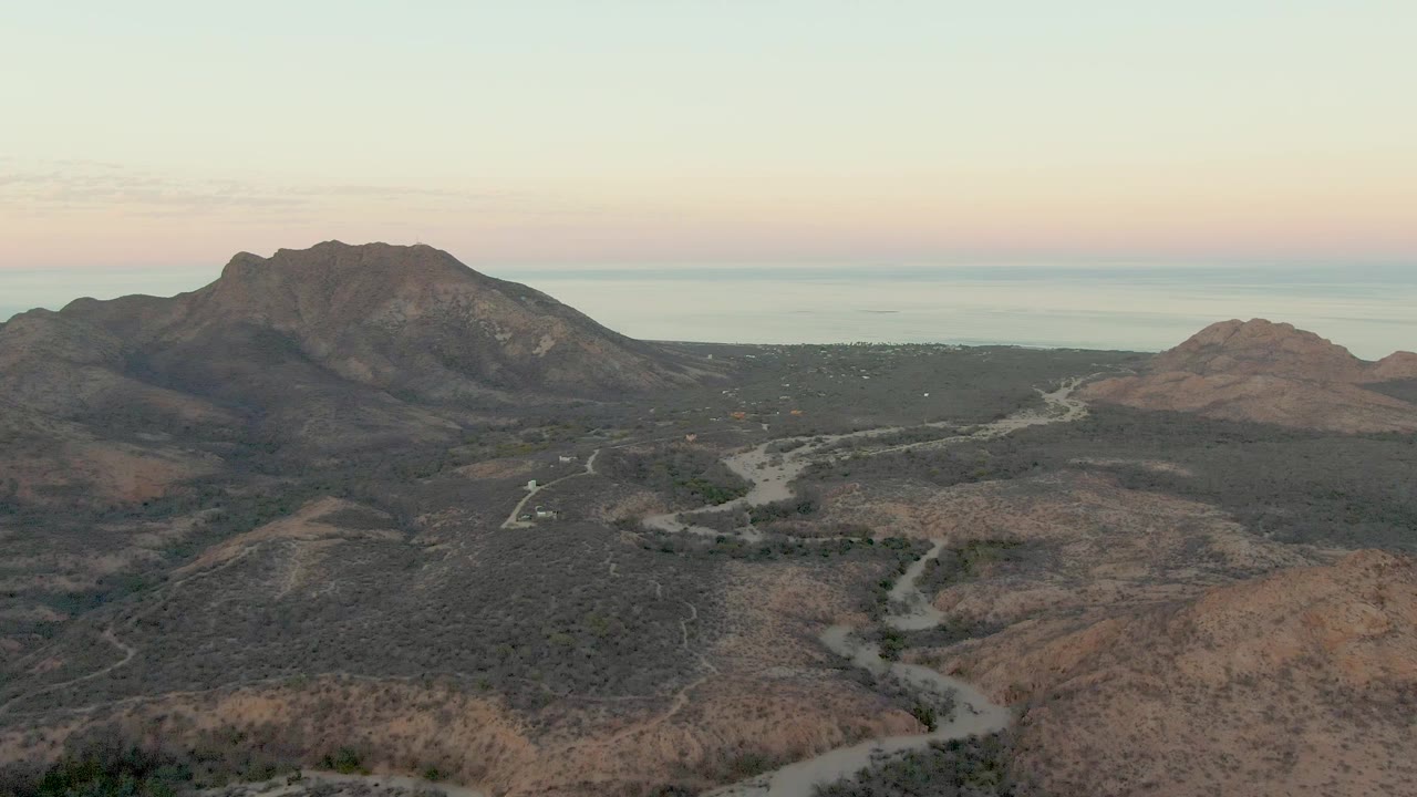 Second frame of the Cabo Pulmo aerial shows branching pale roads across dark scrub and dry hills, with the mountain ridge centered under a softly colored sky and no visible people.