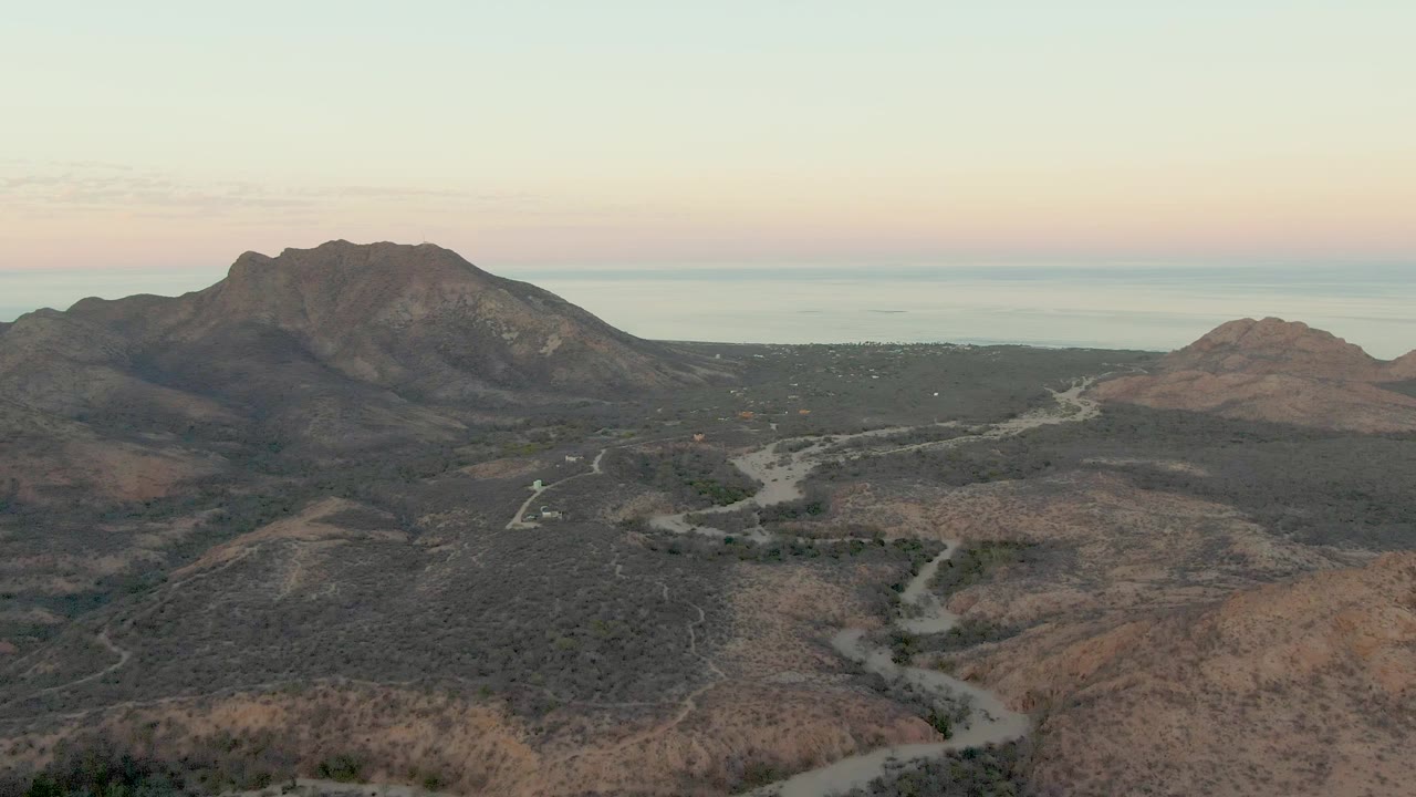 Cabo Pulmo drone view with a pale road curving through the midground, reddish dry hills in front, dark mountain slopes beyond, distant water at the horizon, and no visible people.