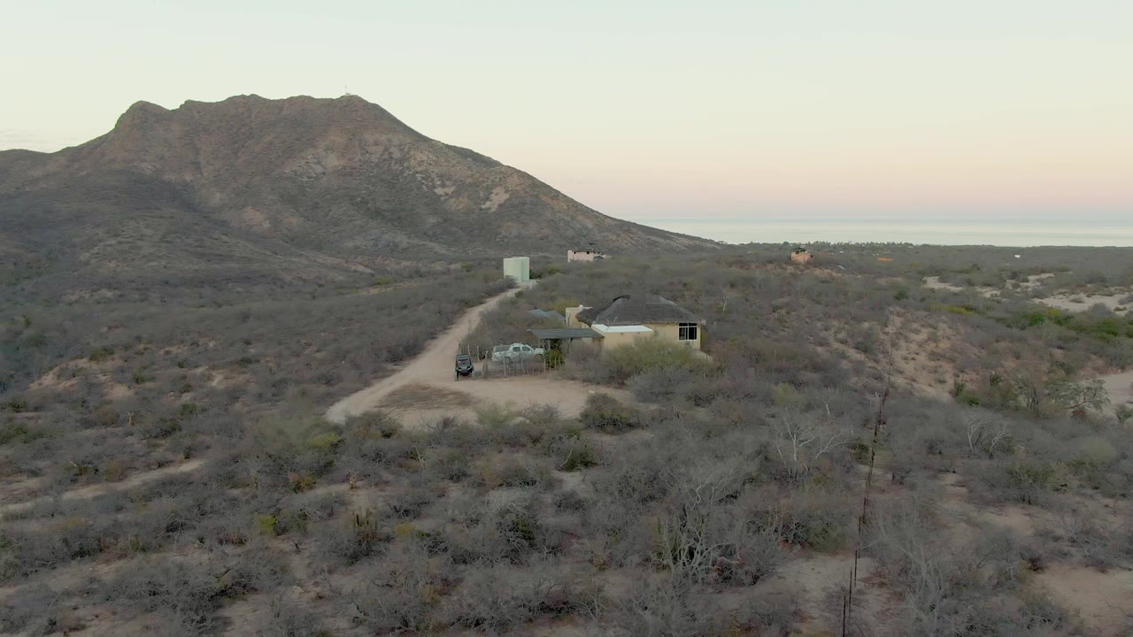 At 0:00, a vehicle is visible on a pale dirt road approaching a small house in the dry Cabo Pulmo desert landscape, with a rocky mountain ridge behind it and no visible people.