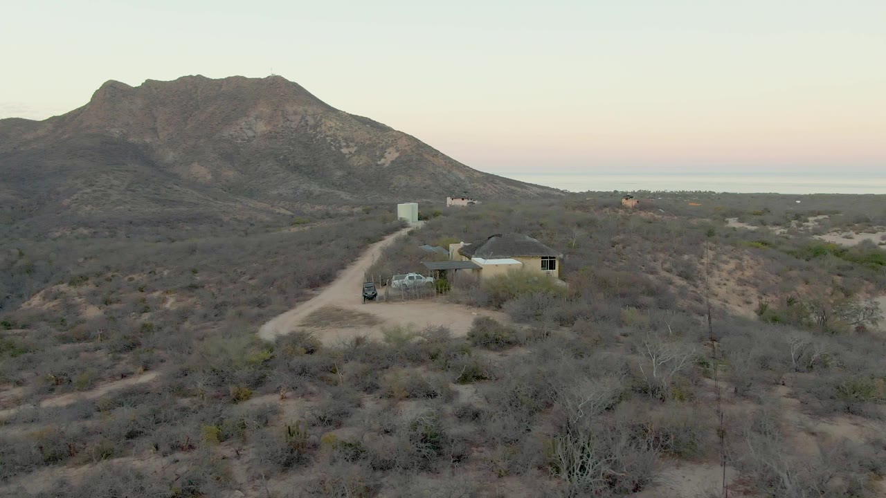 Desert road curving toward a small house beneath a rocky mountain.