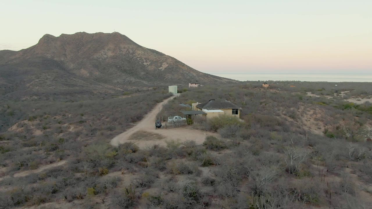 At 0:02, the vehicle remains on the dirt approach road beside the small house, with muted gray-green desert shrubs, a broad rocky mountain, and a faint distant horizon visible in Cabo Pulmo, with no visible people.