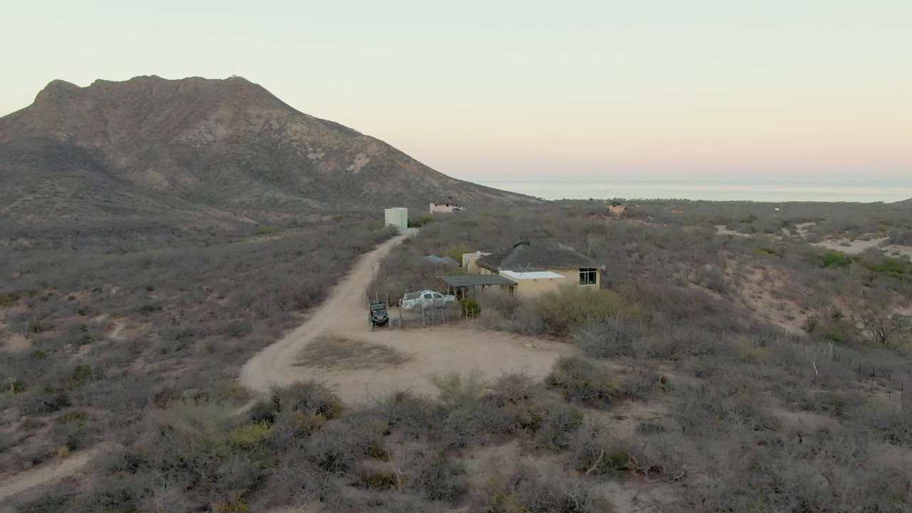 Early aerial frame over Cabo Pulmo desert scrub, with a dirt road leading to a small house and parked vehicle below a rocky mountain, no visible people.