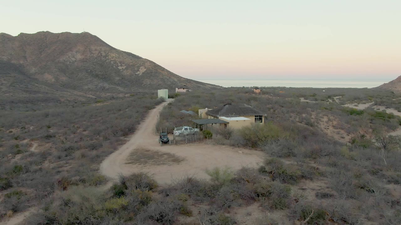 Mid-clip aerial frame in Cabo Pulmo showing dry scrub in the foreground, the dirt road and small house with a parked vehicle in the midground, and pale sky over distant water, no visible people.