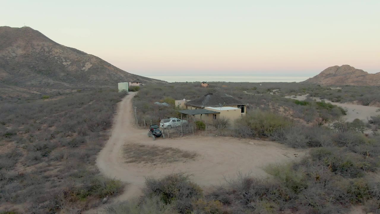 High aerial view in Cabo Pulmo, Baja California Sur, showing a curving dirt road, small desert building, parked light truck, dry scrub, rocky hills, and no visible people.