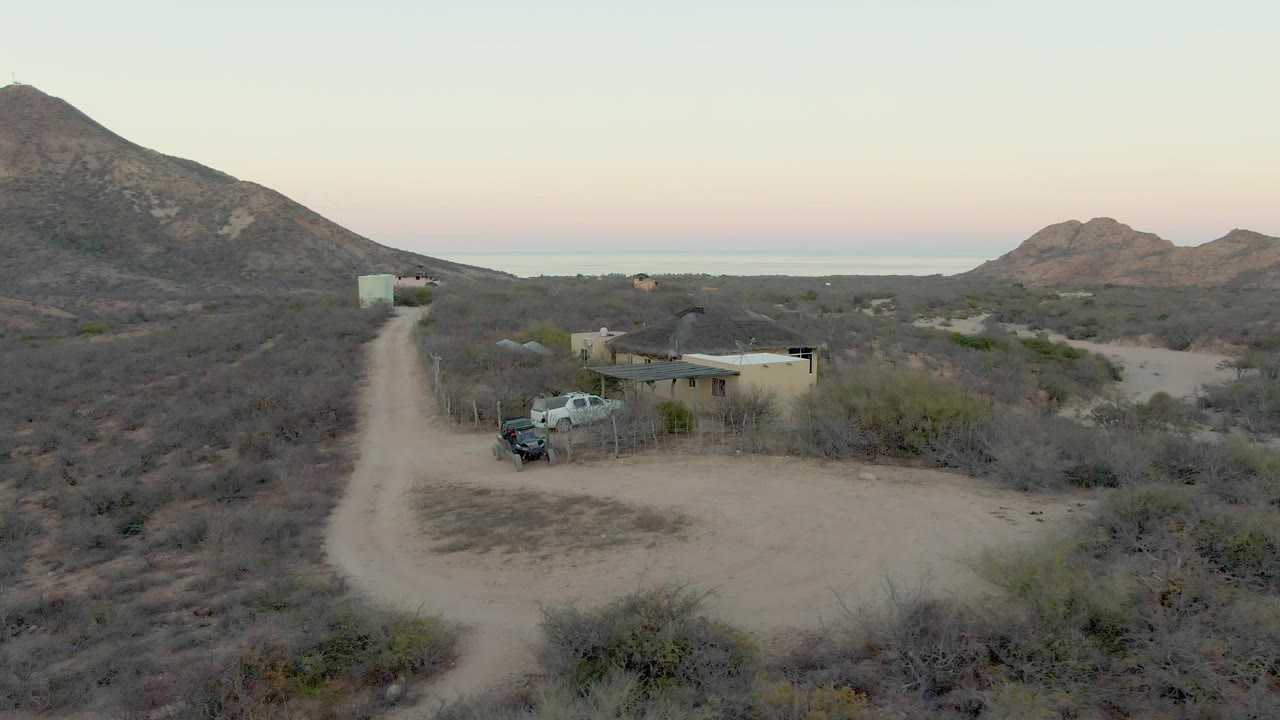 Drone view over the Cabo Pulmo desert property, with the dirt road running from the foreground toward a small building, a parked truck beside it, and no visible people.