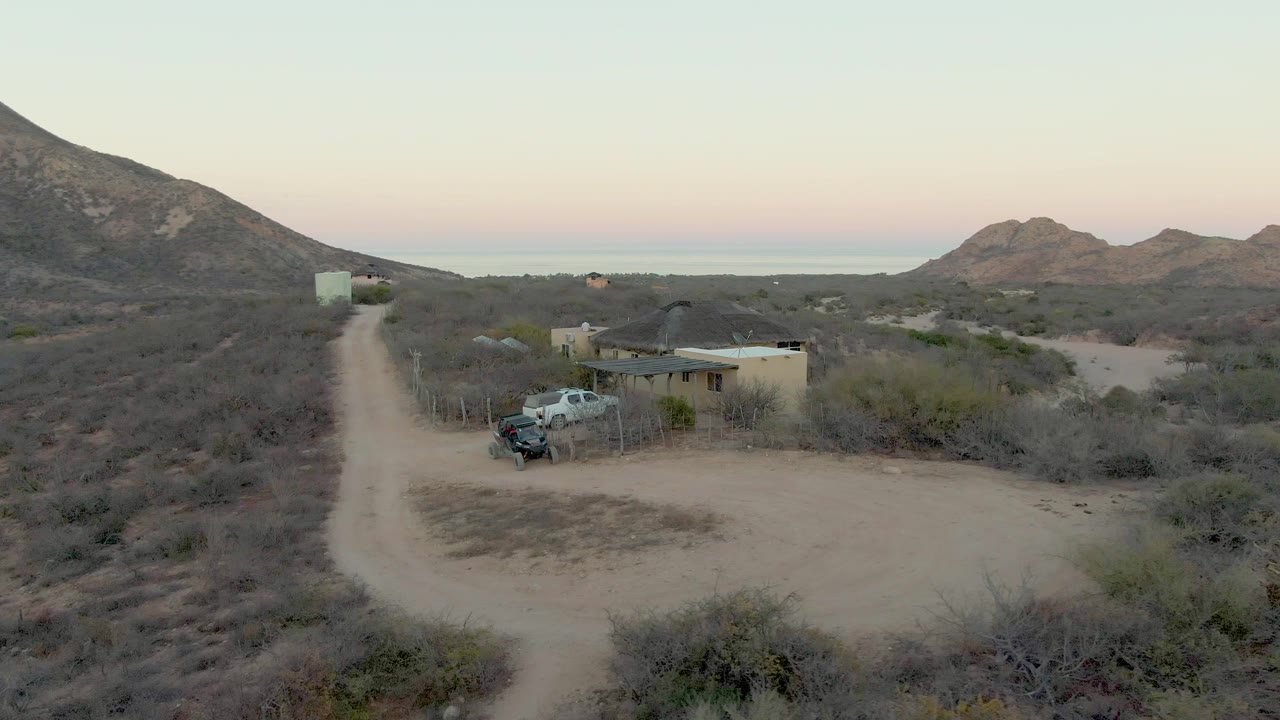 Aerial frame from Cabo Pulmo showing the pale dirt road, low desert structure, light-colored parked truck, surrounding brush, broad hills, and no visible people.