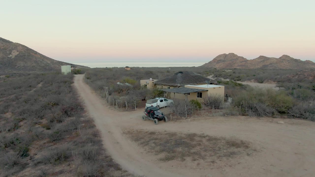 Aerial view in Cabo Pulmo, Baja California Sur, showing a white truck on a dirt road beside dry brush, a small tan house, a parked off-road vehicle, distant mountains, and no visible people.