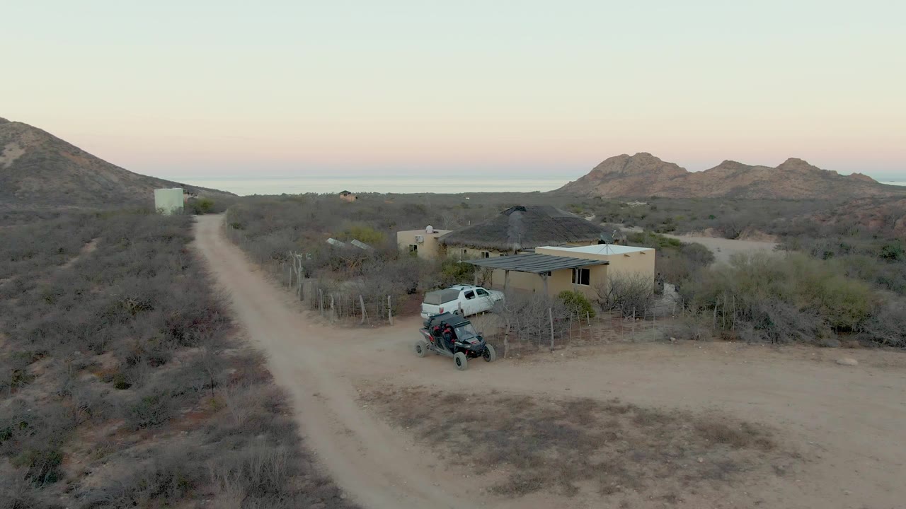 White truck moves closer to a small house beside a dusty road.