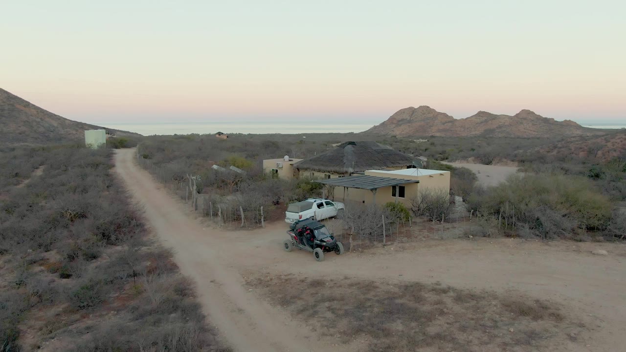Aerial view in Cabo Pulmo with the white truck near the house compound, a dark parked off-road vehicle in front, scrubland surrounding the road, distant rocky hills, and no visible people.