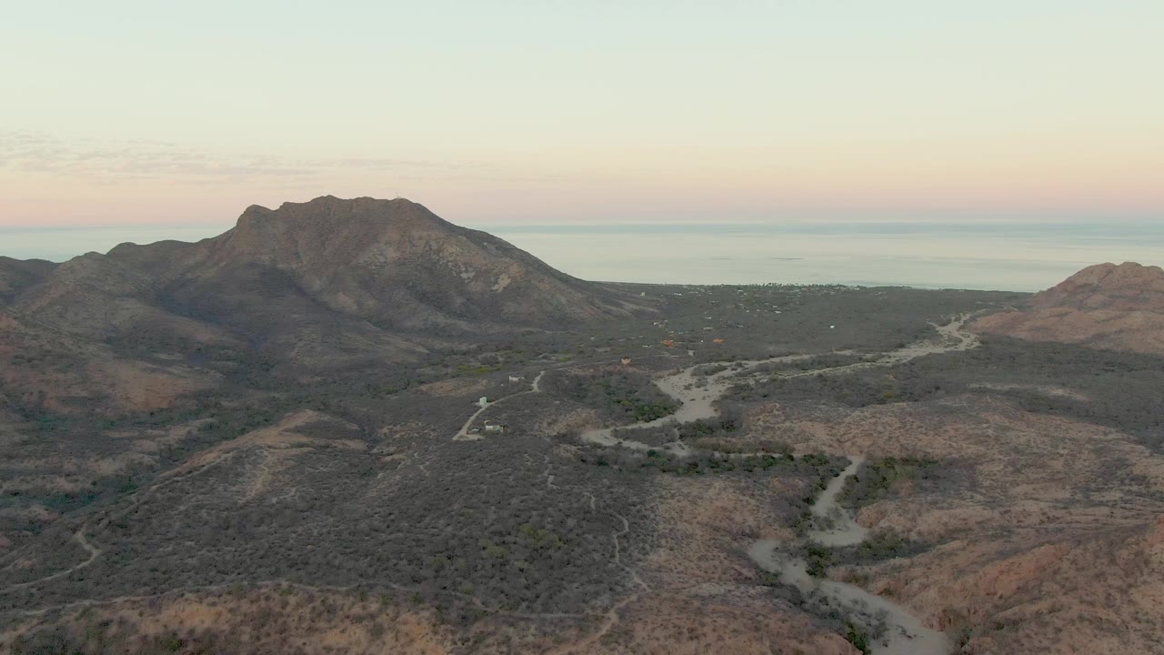 Aerial view of a winding dirt road through dry hills below a rocky mountain and distant ocean.