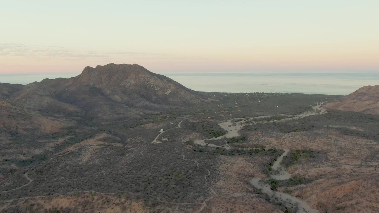 One second into the Cabo Pulmo aerial shot, the pale road bends through scrub-covered hills while the rocky mountain sits center frame and the ocean remains visible behind it, with no visible people.
