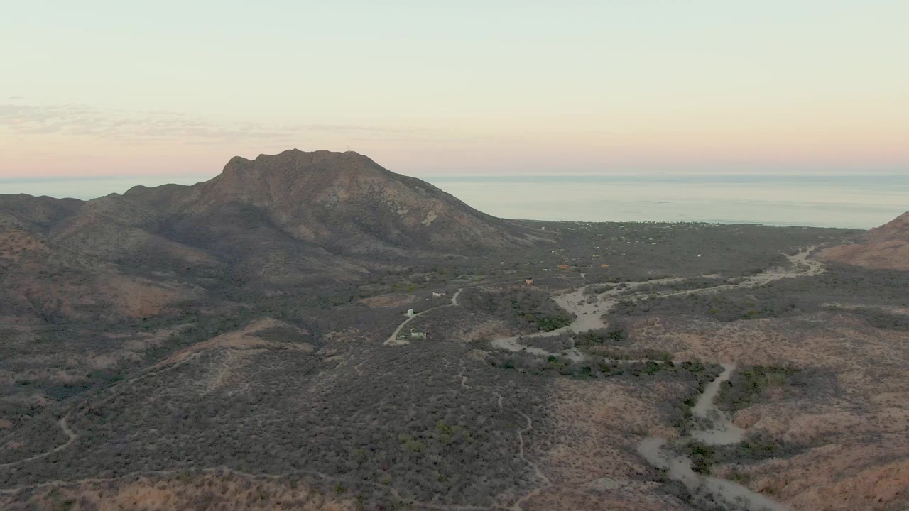 At 0:03, the aerial composition over Cabo Pulmo keeps the winding road low in frame, with scrubby hills rising to the rocky mountain and a thin ocean band behind it, with no visible people.