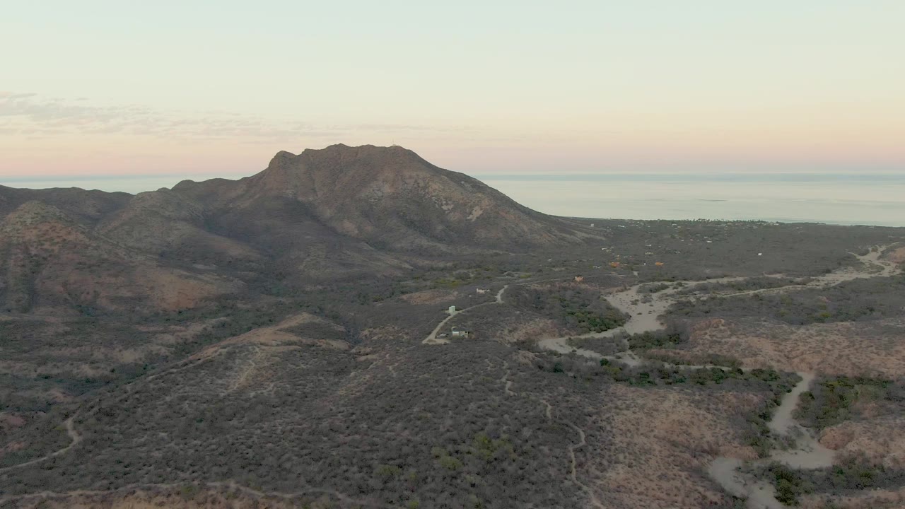 Aerial view of dry hills, winding dirt roads, mountains, and distant sea under a pale sky.